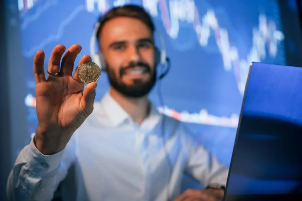 Ein Mann zeigt eine Bitcoin-Münze und sitzt am Laptop