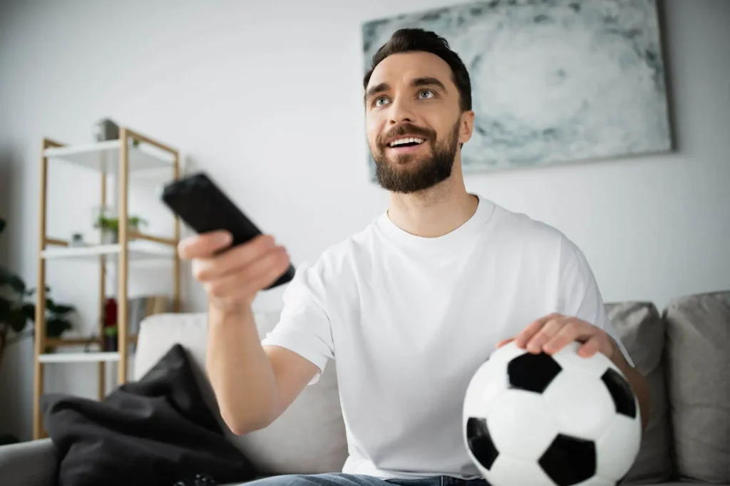 Mann mit Fernbedienung sitzt vor dem TV mit Ball in der Hand