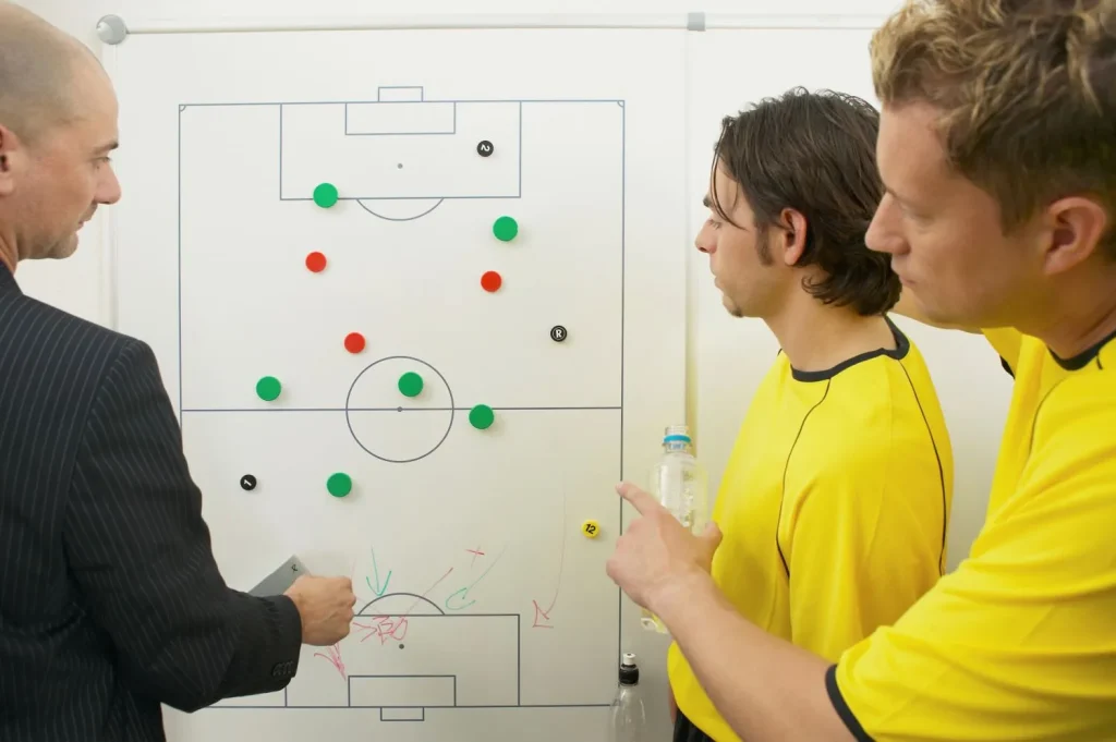 Spieler mit Trainer stehen vor einer Taktiktafel und diskutieren