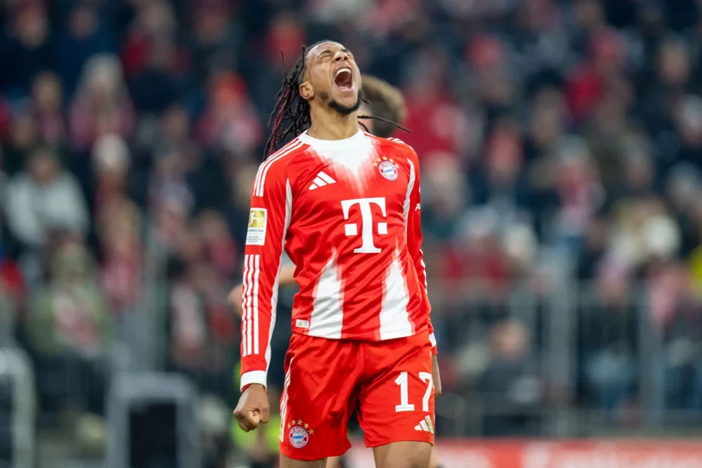Fußballspieler Michael Olise des FC Bayern in rotem Trikot steht während eines Heimspiels im Stadion und jubelt